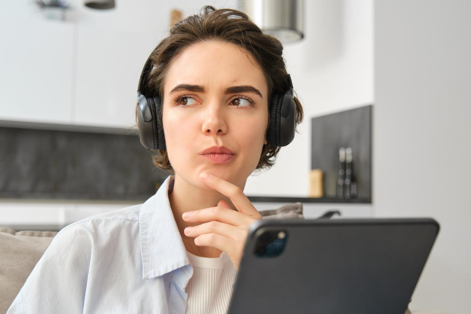 Woman wearing large over-ear headphones rests her chin on her hand and looks to the side, with a tablet in the foreground.