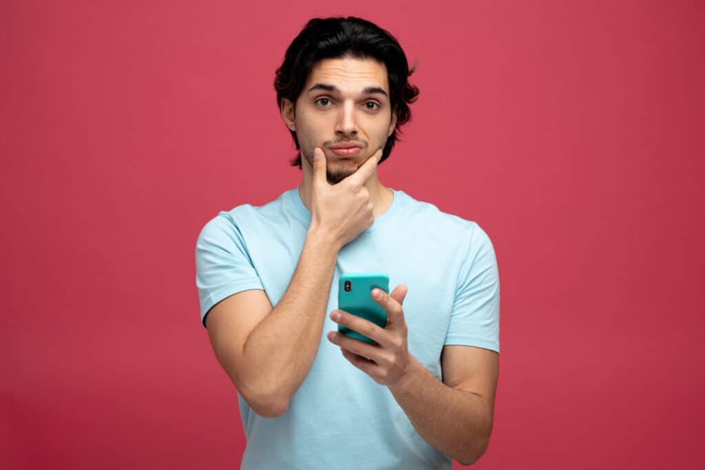 Young man with dark hair in a light blue shirt holds a teal smartphone, thinking with his hand on his chin.