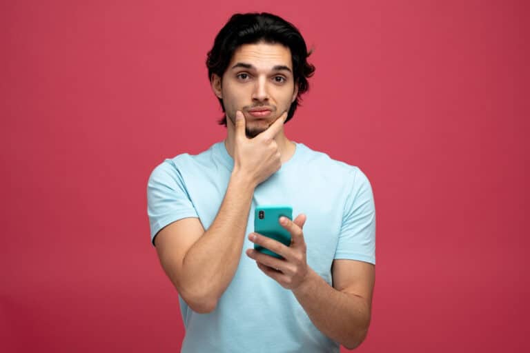 Young man with dark hair in a light blue shirt holds a teal smartphone, thinking with his hand on his chin.