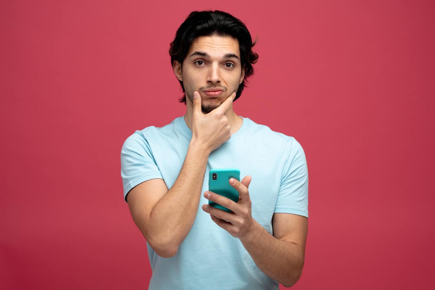 Young man with dark hair in a light blue shirt holds a teal smartphone, thinking with his hand on his chin.
