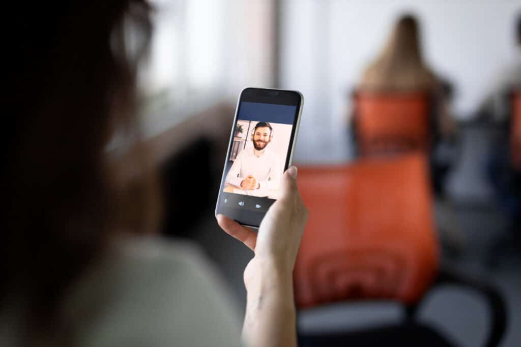 Hand holds a smartphone showing a video call with a bearded man in a white shirt; blurred orange chairs in the background.