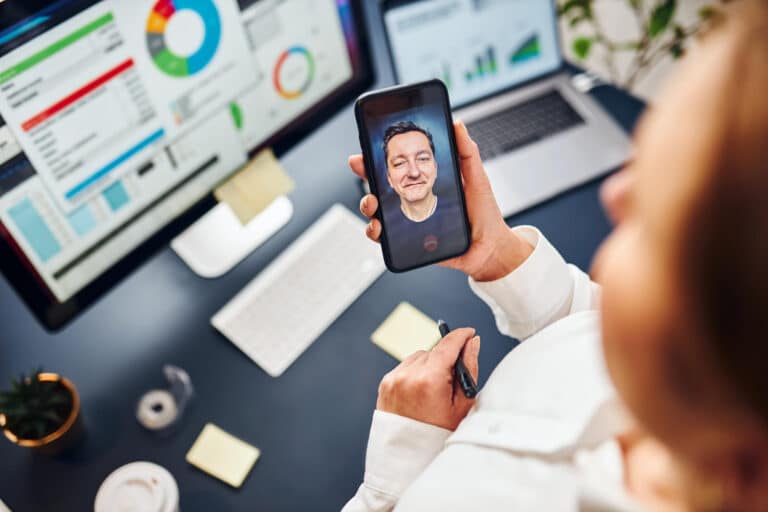 Person holding a phone for a video call with a smiling man; office desk with charts on monitors in the background.