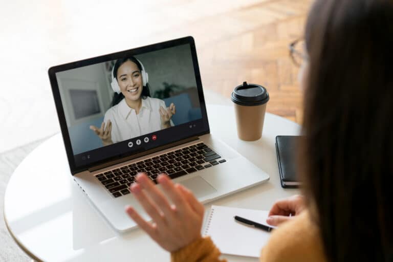 Smiling woman with white headphones on a laptop screen during a video call, with a coffee cup and notebook on the desk.