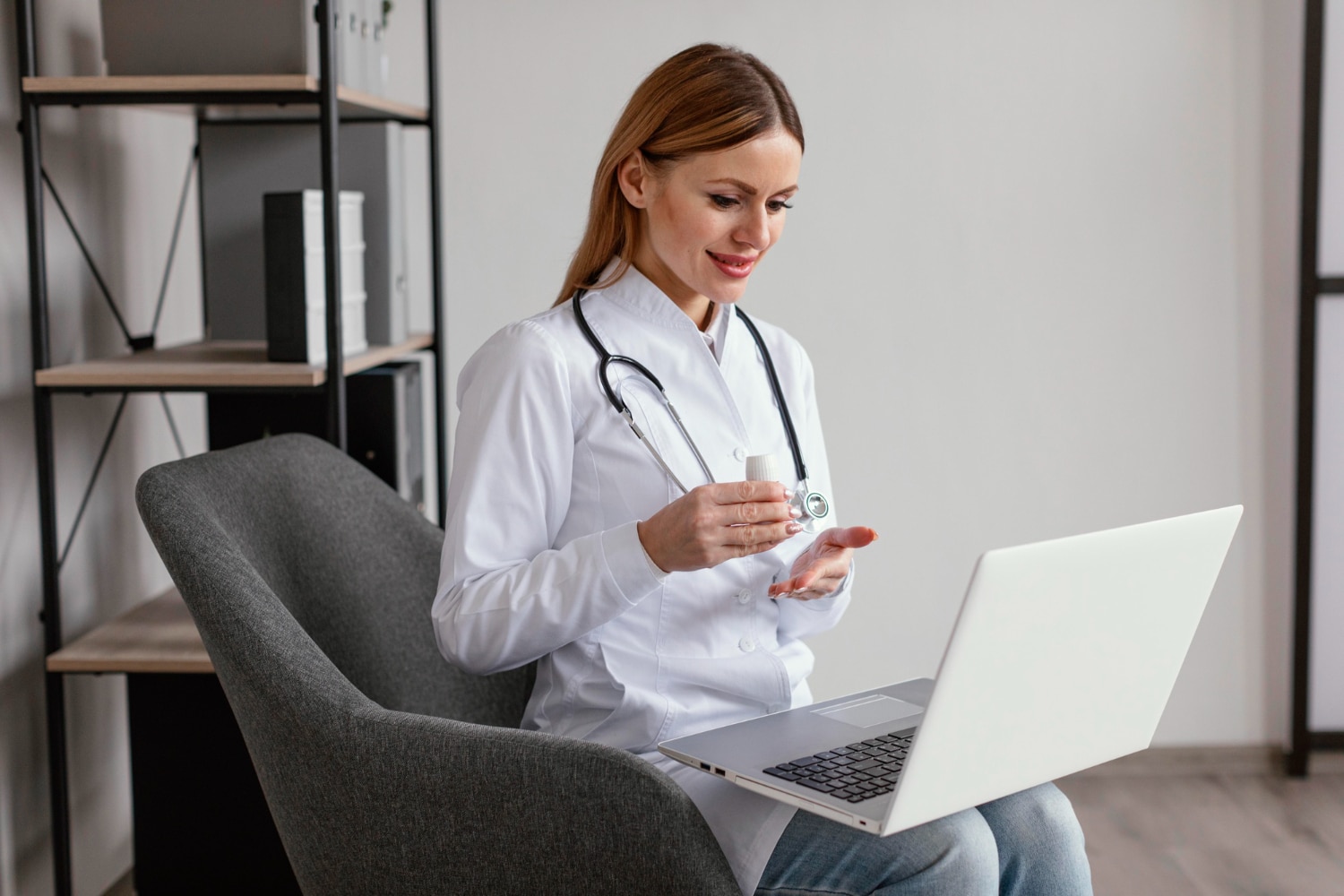 A medical professional in a white coat with a stethoscope around her neck sits in a gray chair, looking at a laptop while holding a small bottle in one hand.