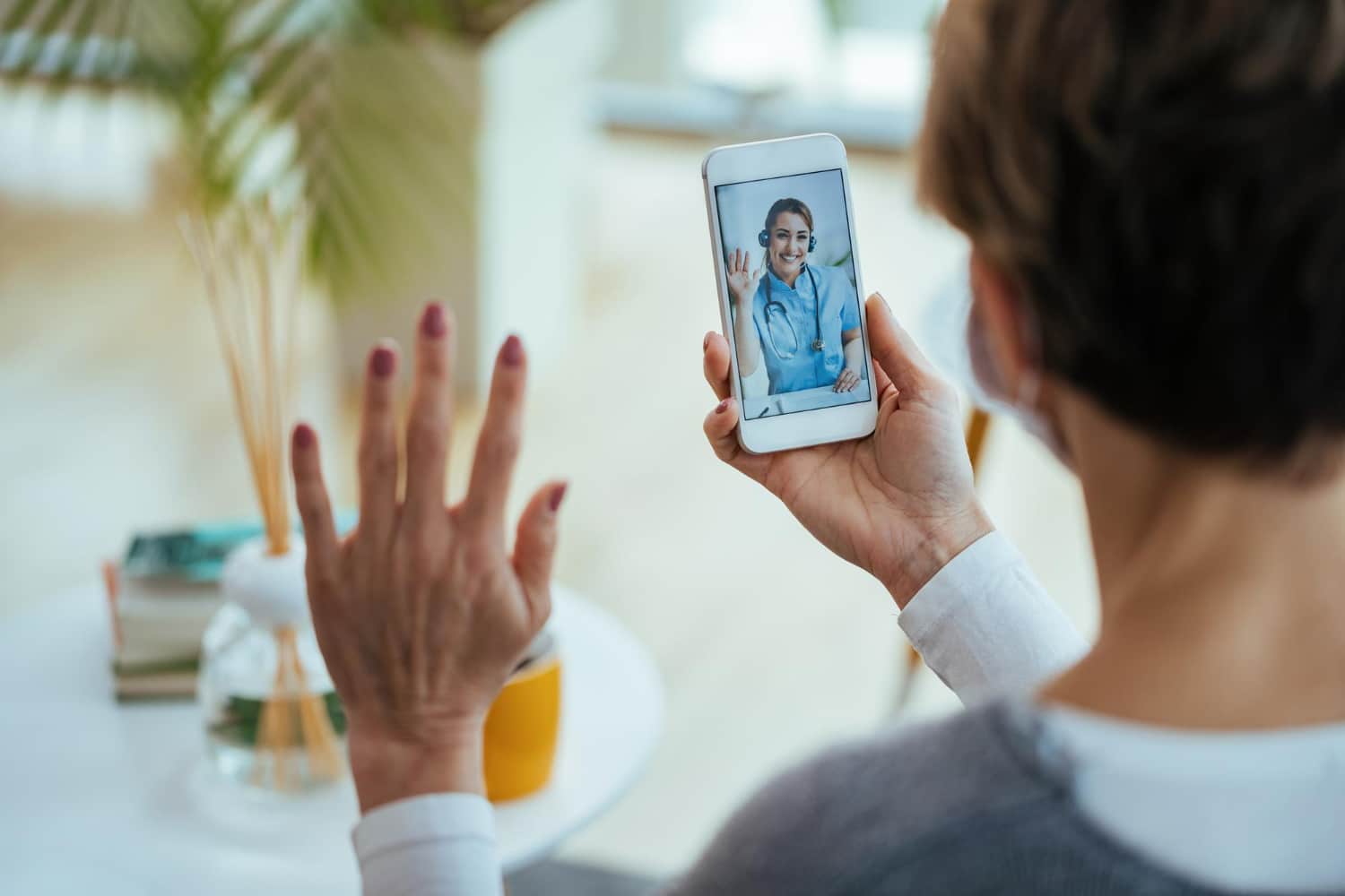 A person video-calling a smiling doctor on a smartphone, waving hello with their other hand raised in view.