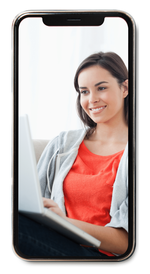 Smiling woman using a laptop while sitting on a couch, wearing a red shirt and gray hoodie.