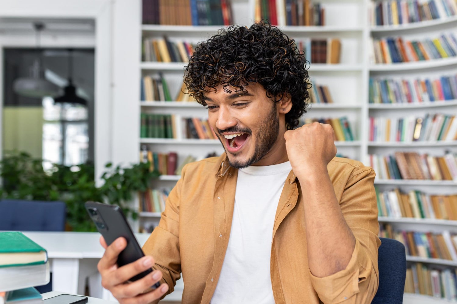 A young man with curly hair and a beard sits at a desk in a library or study area, looking at his phone with excitement, fist clenched in triumph.