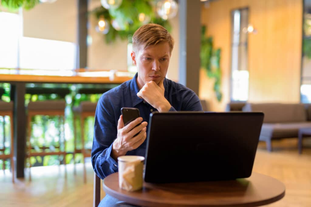 A man in a blue shirt sits at a round wooden table in a bright cafe, looking intently at his smartphone while resting his chin on his hand; a laptop and a coffee cup are nearby.