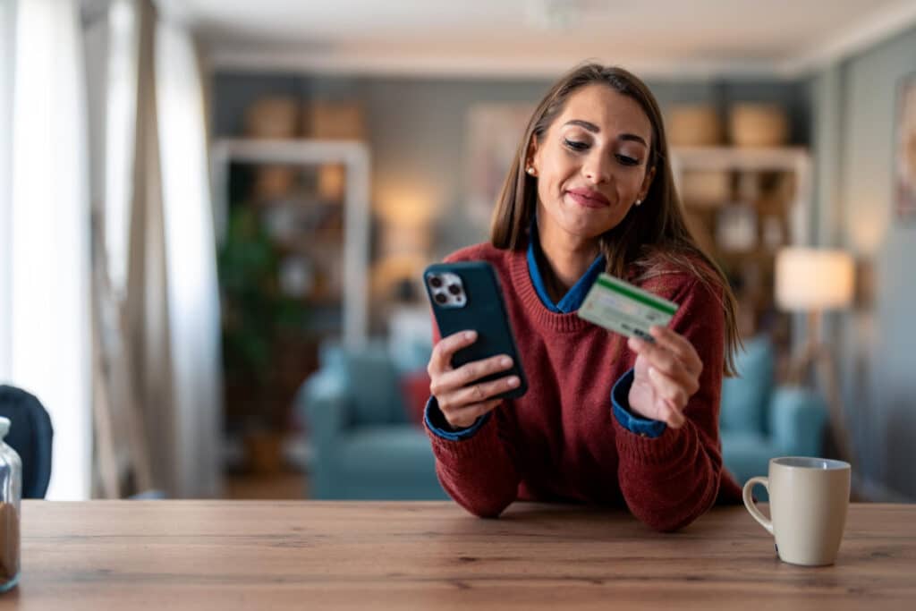 A woman in a red sweater sits at a wooden table, holding a smartphone in one hand and a credit card in the other, smiling as she looks at the phone.