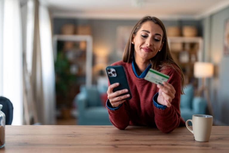 A woman in a red sweater sits at a wooden table, holding a smartphone in one hand and a credit card in the other, smiling as she looks at the phone.