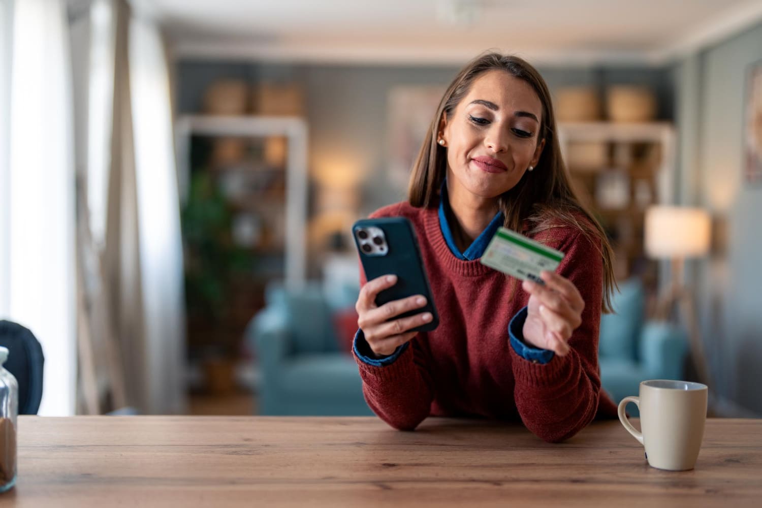 A woman in a red sweater sits at a wooden table, holding a smartphone in one hand and a credit card in the other, smiling as she looks at the phone.