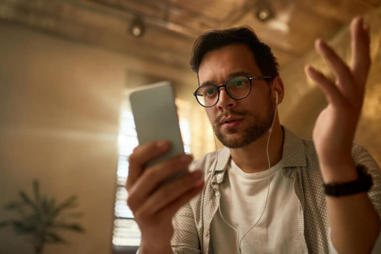 A man with glasses and a beard wearing a white T-shirt and patterned shirt, holding a smartphone in one hand while wearing earphones, looking at the screen with a raised hand gesture in a warm, softly lit room.