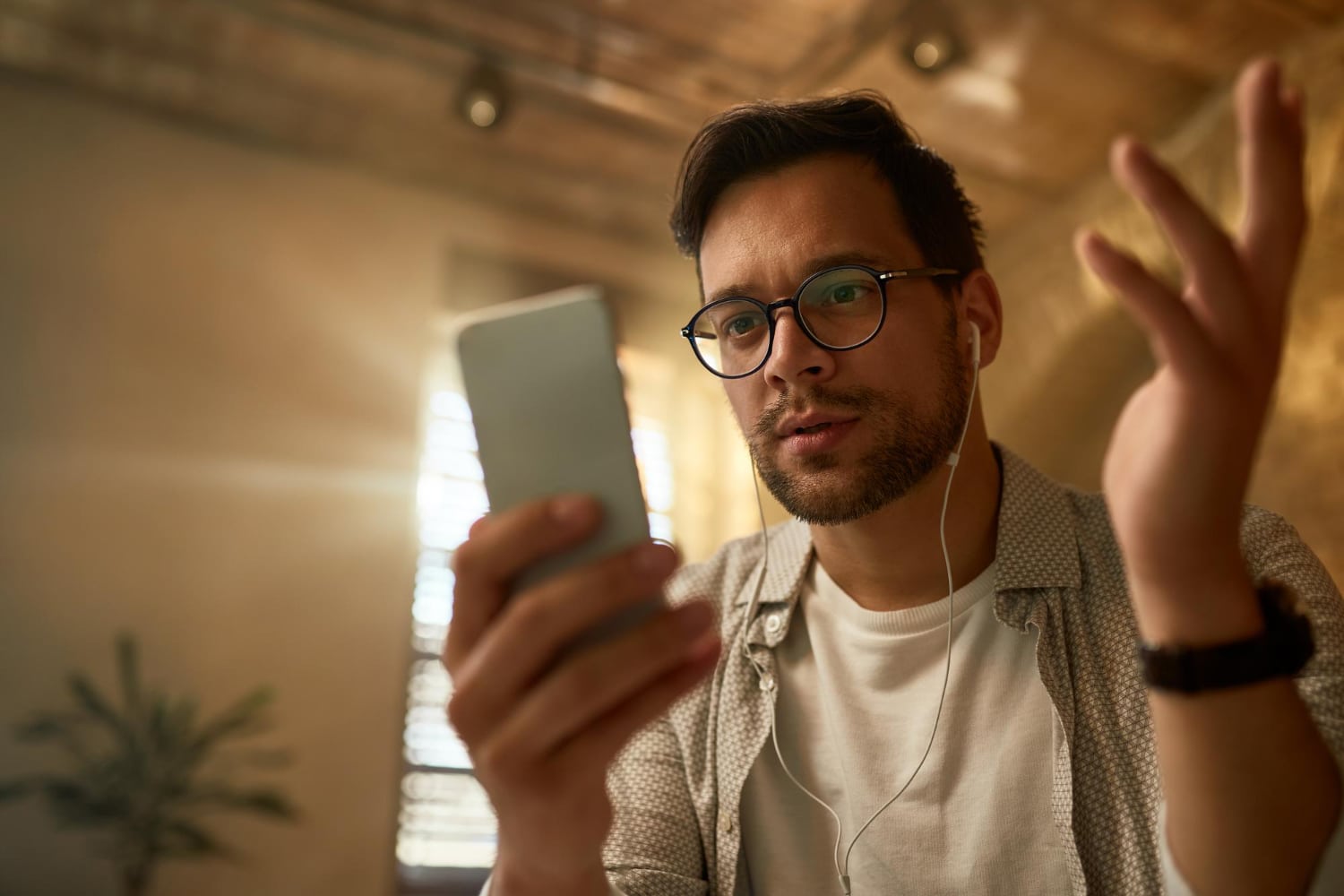 A man with glasses and a beard wearing a white T-shirt and patterned shirt, holding a smartphone in one hand while wearing earphones, looking at the screen with a raised hand gesture in a warm, softly lit room.