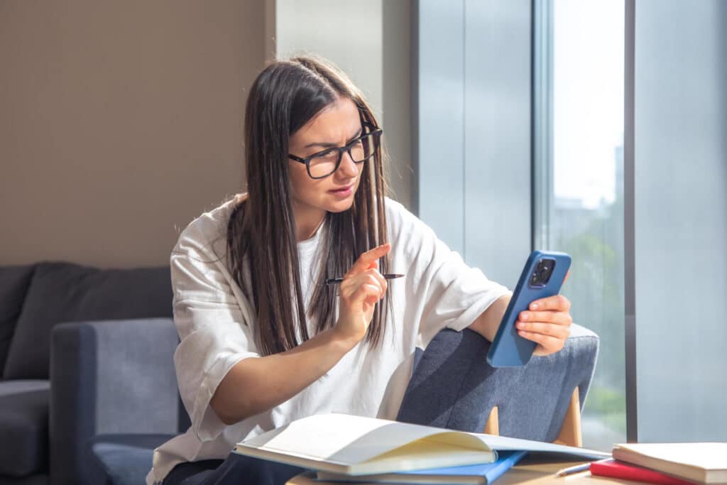 Woman with glasses pointing at a smartphone while studying at a table by a sunlit window