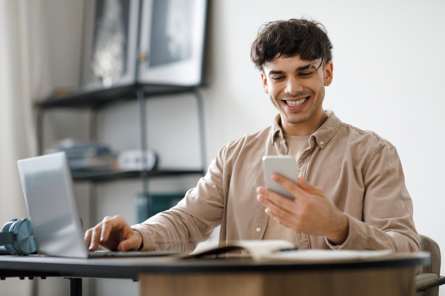 A young man with dark curly hair smiling while looking at his smartphone, sitting at a desk with a laptop and headphones