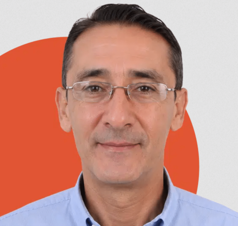 A man with short dark hair, wearing glasses and a light blue shirt, smiling in front of a background with a red-orange circle on a light gray backdrop