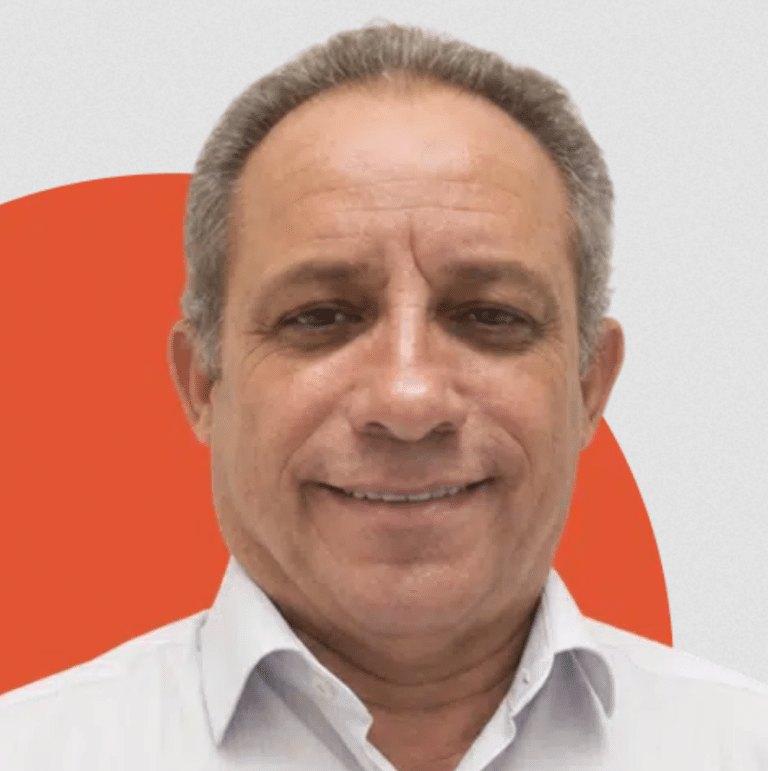 A smiling middle-aged man with short gray hair wearing a white collared shirt, standing in front of a background with orange and white shapes