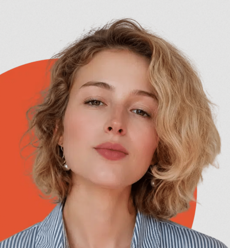 A young woman with wavy blonde hair, fair skin, light makeup, wearing earrings, and a striped shirt, posing against a background with orange and white areas